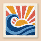 A square punch needle or tufted art piece on a cream fabric background, featuring a stylized ocean wave and rising sun in blue, navy, yellow, orange, and white, shown against a light beige backdrop.